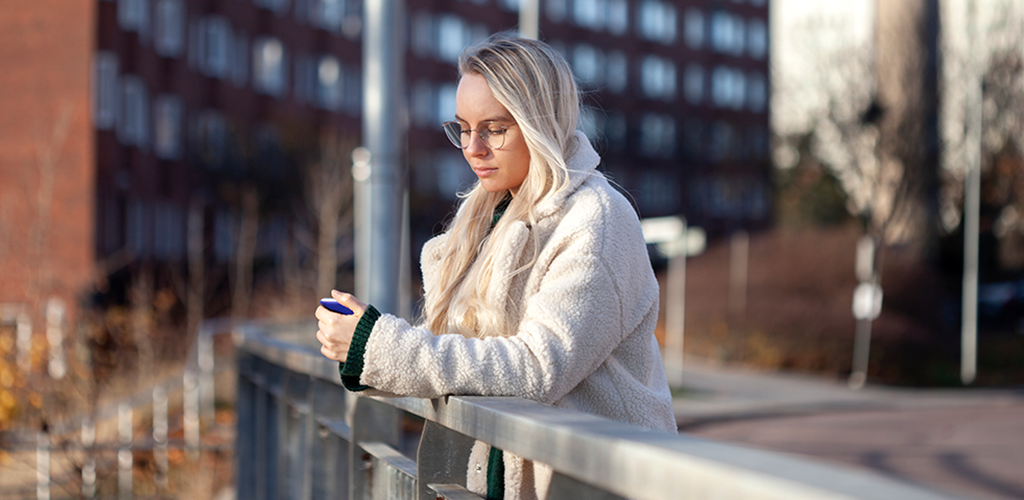 Ung kvinna står och tittar på sin mobil med lägenheter i bakgrunden.