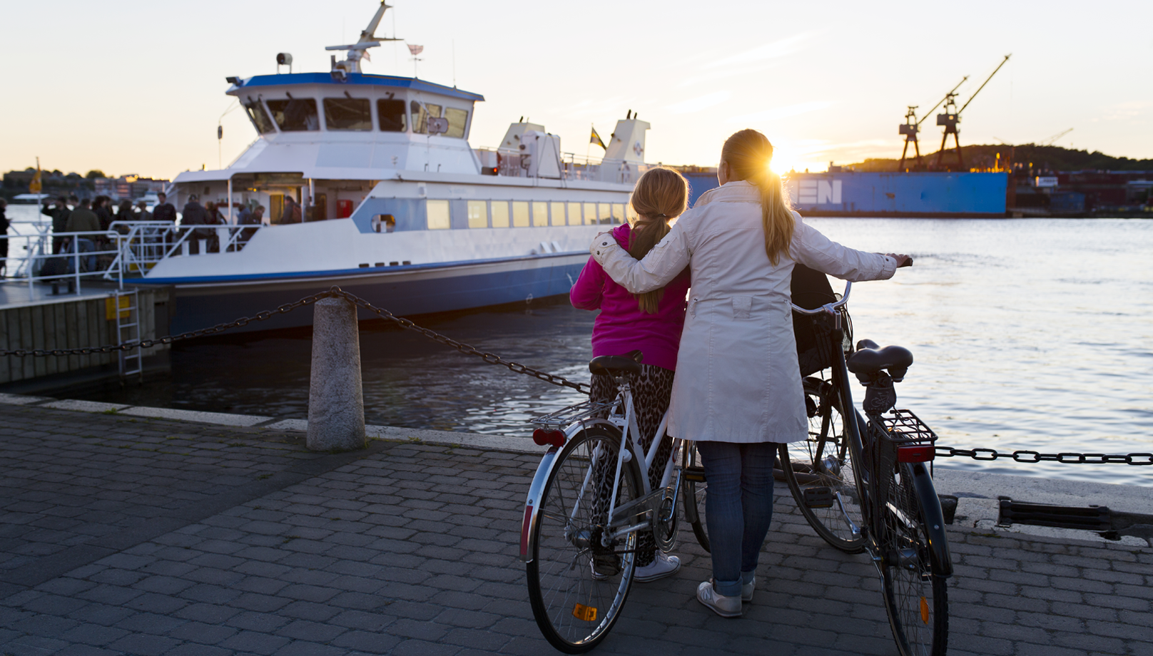 En kvinna och ett barn står med två cyklar vid havet. I havet ligger en kollektivtrafiksbåt.
